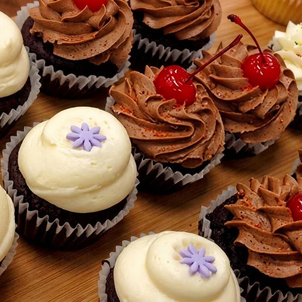 Assortment of chocolate cupcakes with white or chocolate frosting, topped with cherries or purple flowers.