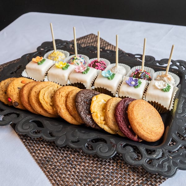 A black ornate tray holds a variety of colorful cookies, petit fours, and cake pops.