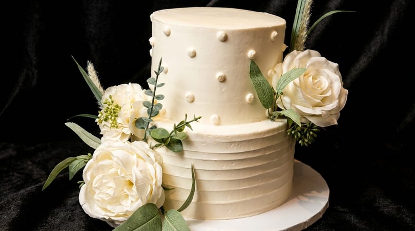 Two-tier white cake decorated with white roses, green foliage, and textured frosting on a dark background.