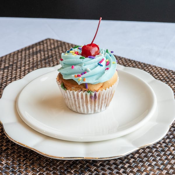 A vanilla cupcake with light blue frosting, colorful sprinkles, and a cherry on top, on white plates.