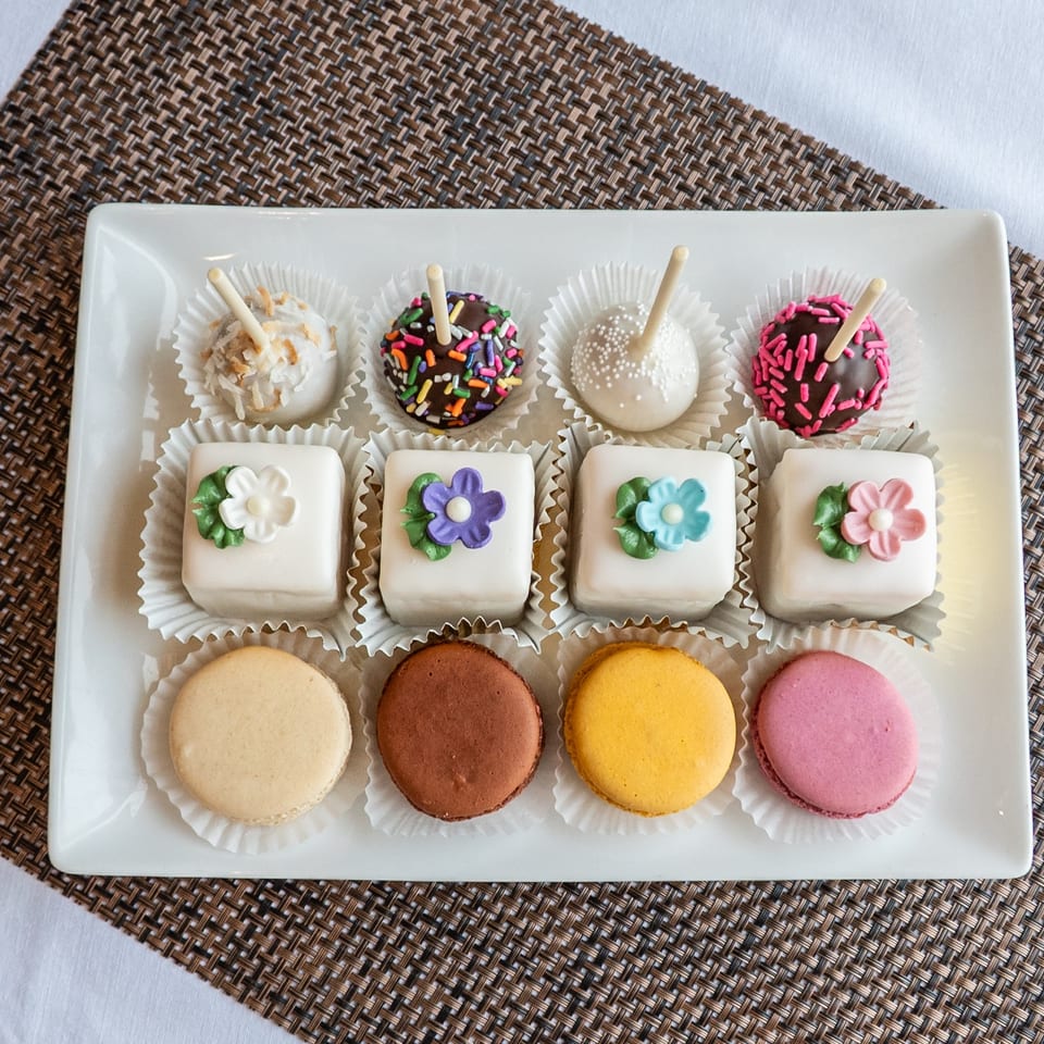 Assorted decorated cake pops, flower-topped petit fours, and colorful macarons arranged on a white platter.