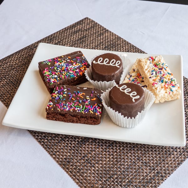 A white rectangular plate holds two brownies, two chocolate cakes, and two rice crispy treats, all with colorful sprinkles.