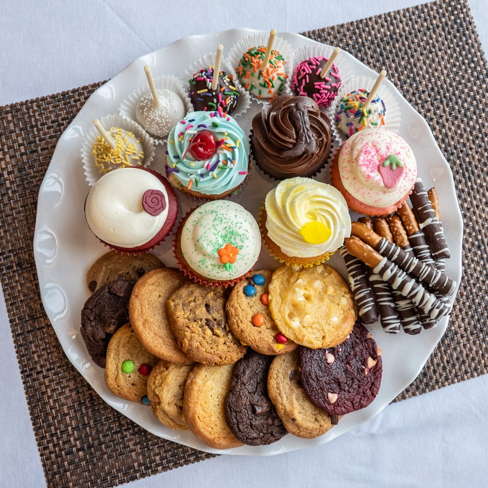 A white platter filled with an assortment of colorful cupcakes, cake pops, cookies, and chocolate-dipped pretzels.