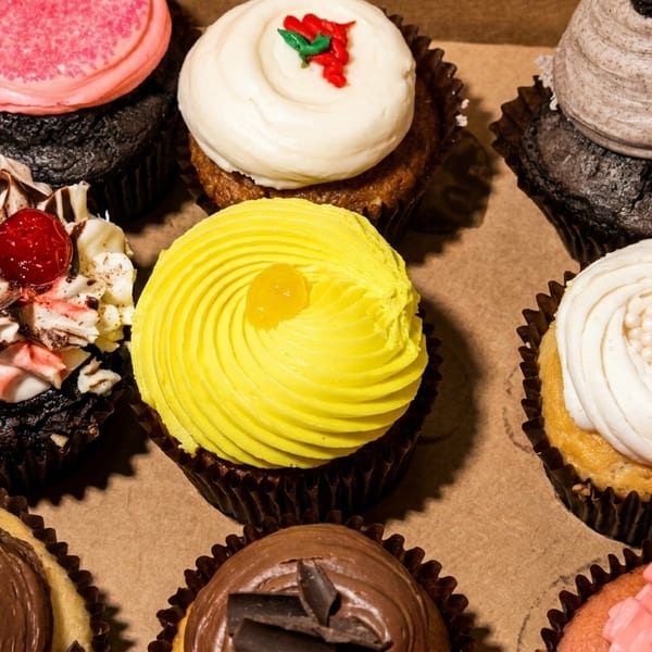 Assortment of nine colorful cupcakes with various frostings and toppings in a brown box.